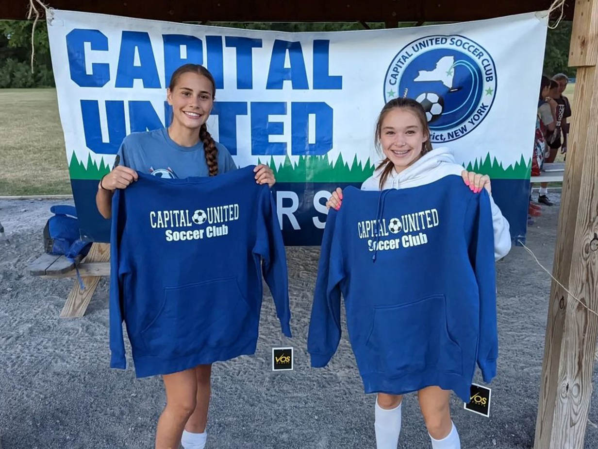 Two girls soccer players holding up Capital Elite FC hooded sweatshirts.